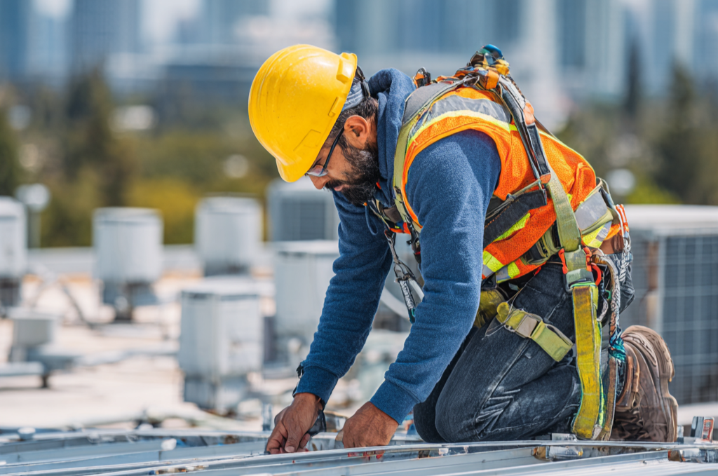 Hispanic roofer in full PPE including hard hat, hi-vis vest, and safety harness working on commercial construction site rooftop with city skyline background